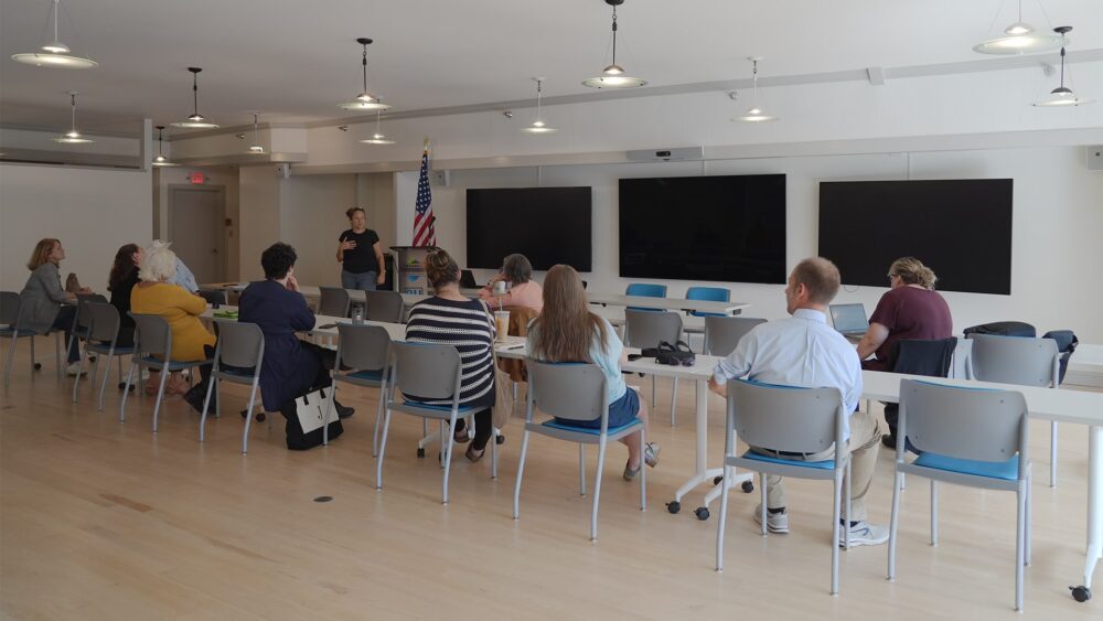 Photo of the U.S. Committee for Refugees and Immigrants (USCRI) meeting at The Hub CoWorks in Rutland, Vermont, on October 1, 2024. Attendees, including local community partners, are shown engaging in a discussion about USCRI’s accomplishments, plans for 2025, and ongoing collaboration to support refugees and immigrants. The Streetside Conference Room is filled with participants actively involved in the conversation.