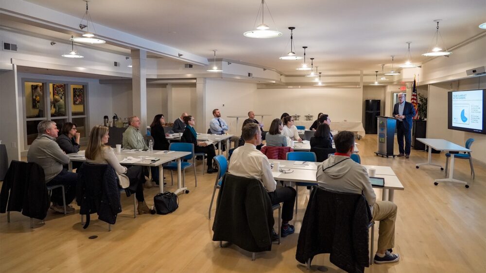 Attendees at the Rutland Regional Medical Center (RRMC) event gather in The Hub CoWorks’ large, tech-enabled Streetside Conference Room in downtown Rutland, Vermont. The modern coworking space features collaborative seating, state-of-the-art Cisco Webex technology for hybrid meetings, and three large screens displaying healthcare data and presentations, including topics like the False Claims Act. The room’s sleek design, natural light, and professional yet welcoming atmosphere make it ideal for events, meetings, and presentations.