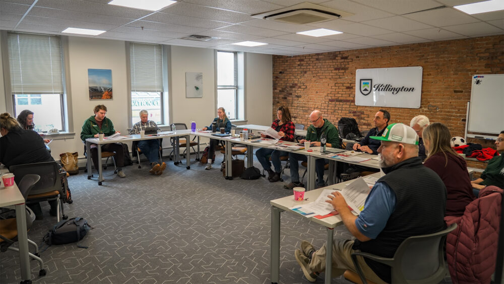 Come Alive Outside's Board Meeting and strategy session in the Killington room at The Hub CoWorks in downtown Rutland, Vermont. Members are sitting at grey tables in the shape of a "U" with a white wall and a brick wall in the background with the Killington logo on the wall.