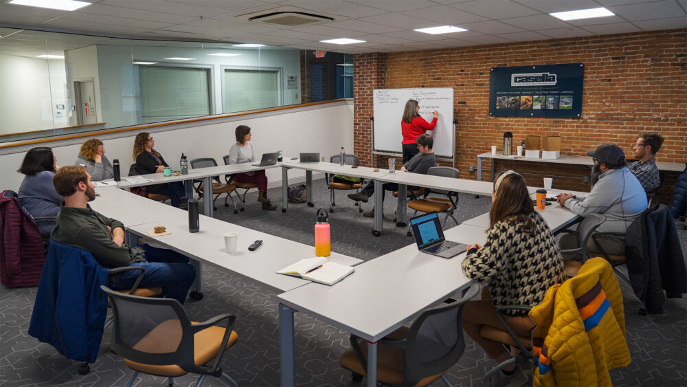 Members of The MINT at a Board Strategy Meeting in a modern coworking space with a group of engaged attendees seated around a U-shaped table setup, actively participating in a presentation by a facilitator at a whiteboard. The space features exposed brick walls, glass partitions, and a banner for Casella in the background, reflecting a collaborative and business-oriented environment.