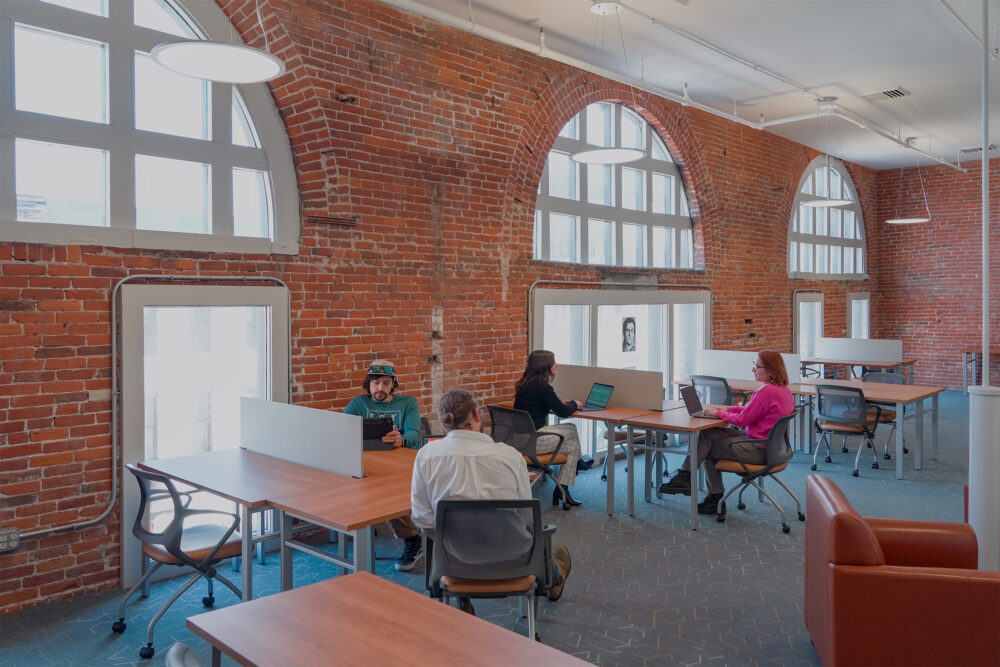 A photograph of people working in a modern coworking space with brick walls and large windows. This space is called the Opera House Lounge at The Hub CoWorks in downtown Rutland, Vermont. Rutland's premiere and best coworking, meeting space, and office space.