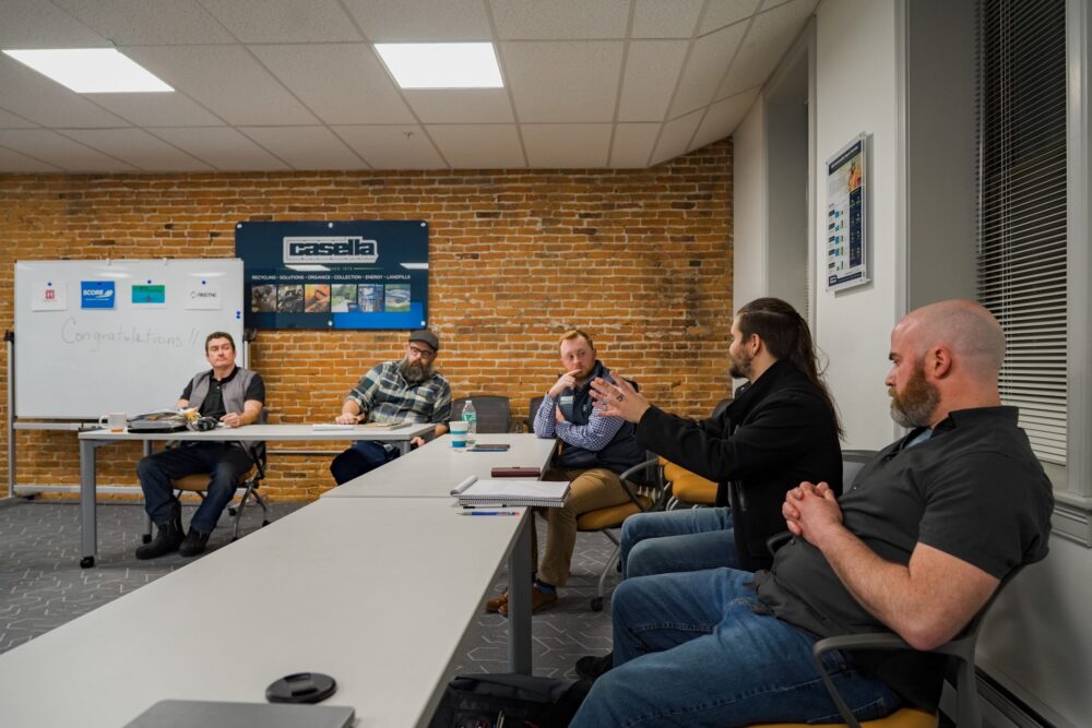 The graduates of the first CO.STARTERS cohort at The Hub CoWorks are seated around a conference table in the Casella Room at The Hub CoWorks. One graduate in a black coat is gesturing while speaking, while the others listen attentively. A whiteboard in the background displays the word “Congratulations!!” alongside logos for SCORE, FINSYNC, and Heritage Family Credit Union. A Casella sign is mounted on the exposed brick wall. The setting captures a moment from their final session, marking the completion of the 10-week business accelerator program.