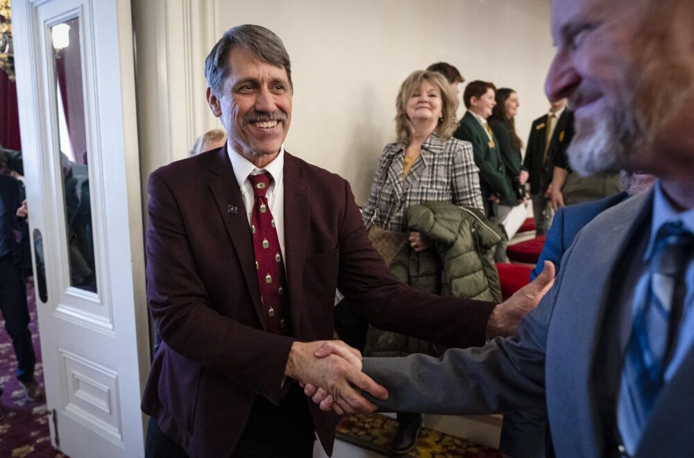 Vermont Lieutenant Governor John Rodgers, wearing a burgundy suit and patterned tie, smiles while shaking hands with a man in a suit at an indoor event. Behind him, a woman in a plaid coat and other attendees, including students in green blazers, observe the interaction. The setting features classic architectural details, including white doors and red-carpeted steps.