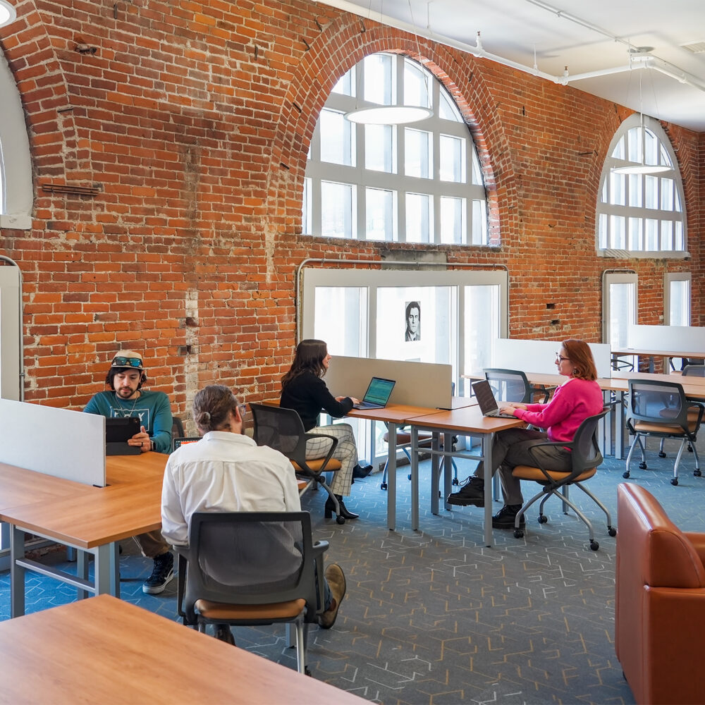 People in a modern coworking space with large windows in a brick wall, letting in a large amount of natural light.