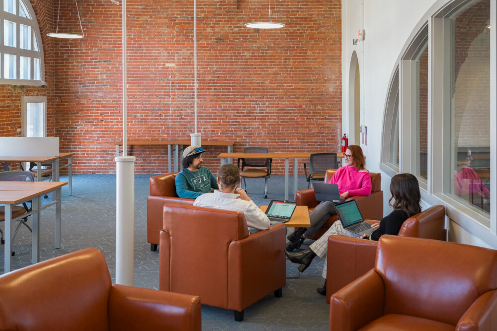Four people sitting in orange chairs around a table in conversation. They are in a coworking space with a brick wall and a lot of natural light in downtown Rutland, Vermont.
